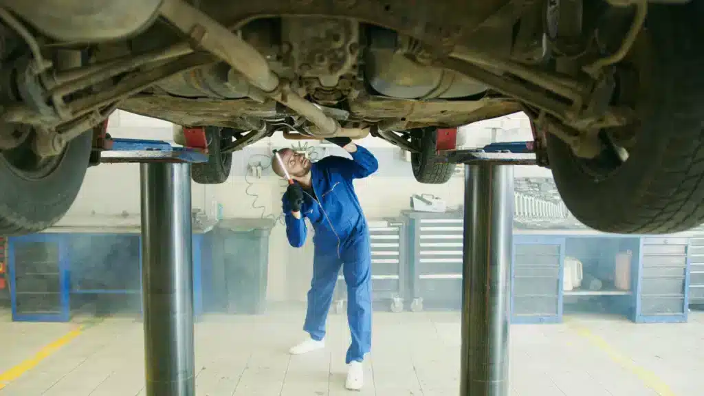 Mechanic in a blue jumpsuit inspecting the underside of a car on a lift in an auto repair workshop.